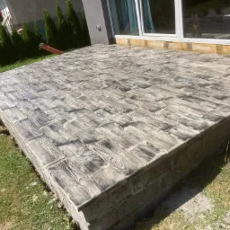 Nowo ułożony, betonowy taras z odciskiem imitującym drewniane deski, otoczony trawnikiem i elementami budynku.