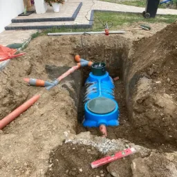 Niebieski zbiornik przydomowej oczyszczalni ściek&oacute;w umieszczony w wykopie, widoczne rury kanalizacyjne i poziomica, w tle dom z kostką brukową i kwiatami.