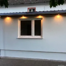 Otynkowana, pomalowana na biało dobudówka z oknem PCV i zewnętrznym oświetleniem punktowym, z widoczną rynną odprowadzającą wodę, przylegająca do starszego budynku z czerwonymi elementami...