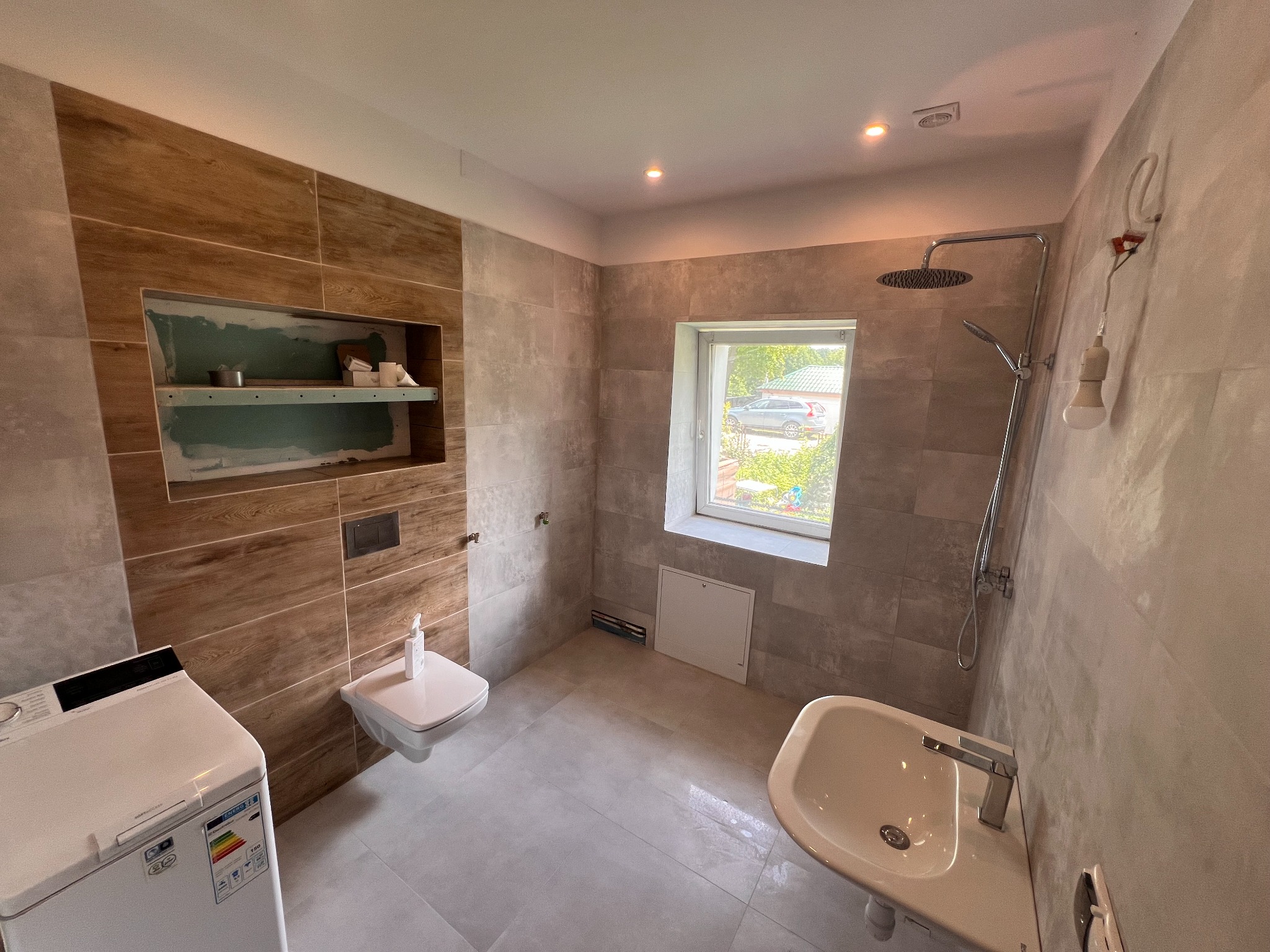 Nowoczesna łazienka w trakcie wykończenia: płytki imitujące drewno, wisząca toaleta, umywalka, pralka i półka wnękowa w ścianie z narzędziami.