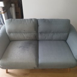 Clean-Pro - Dwuosobowa kanapa w odcieniach szarości, pokazująca różnicę w wyglądzie przed i po czyszczeniu, z wyraźnie widoczną plamą na lewej połowie i czystą prawą połową. Widoczne pudełka w tle i fragment...
