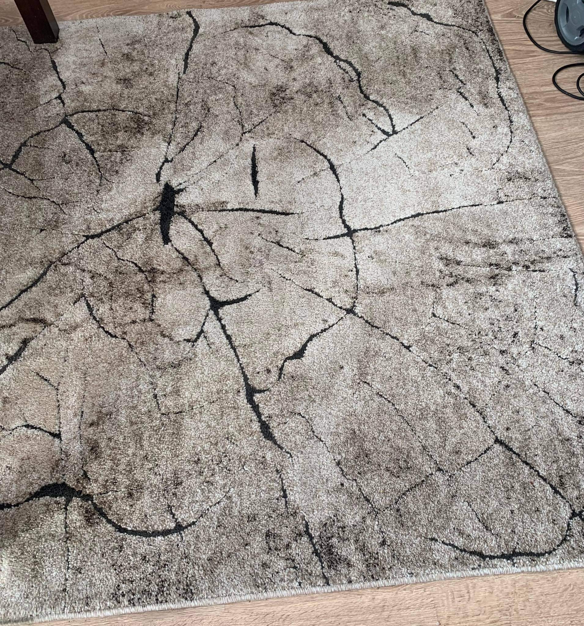Beżowy dywan z abstrakcyjnym wzorem przypominającym pęknięcia kory drzewa, leżący na drewnianej podłodze, z fragmentem nogi meblowej w rogu.