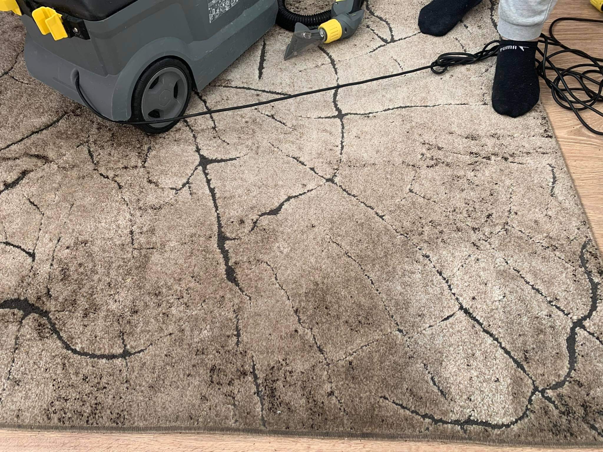 Czyszczenie beżowego dywanu z ciemnymi wzorami za pomocą szarego odkurzacza piorącego z żółtymi elementami; widoczne stopy w czarnych skarpetach Puma i szare spodnie.