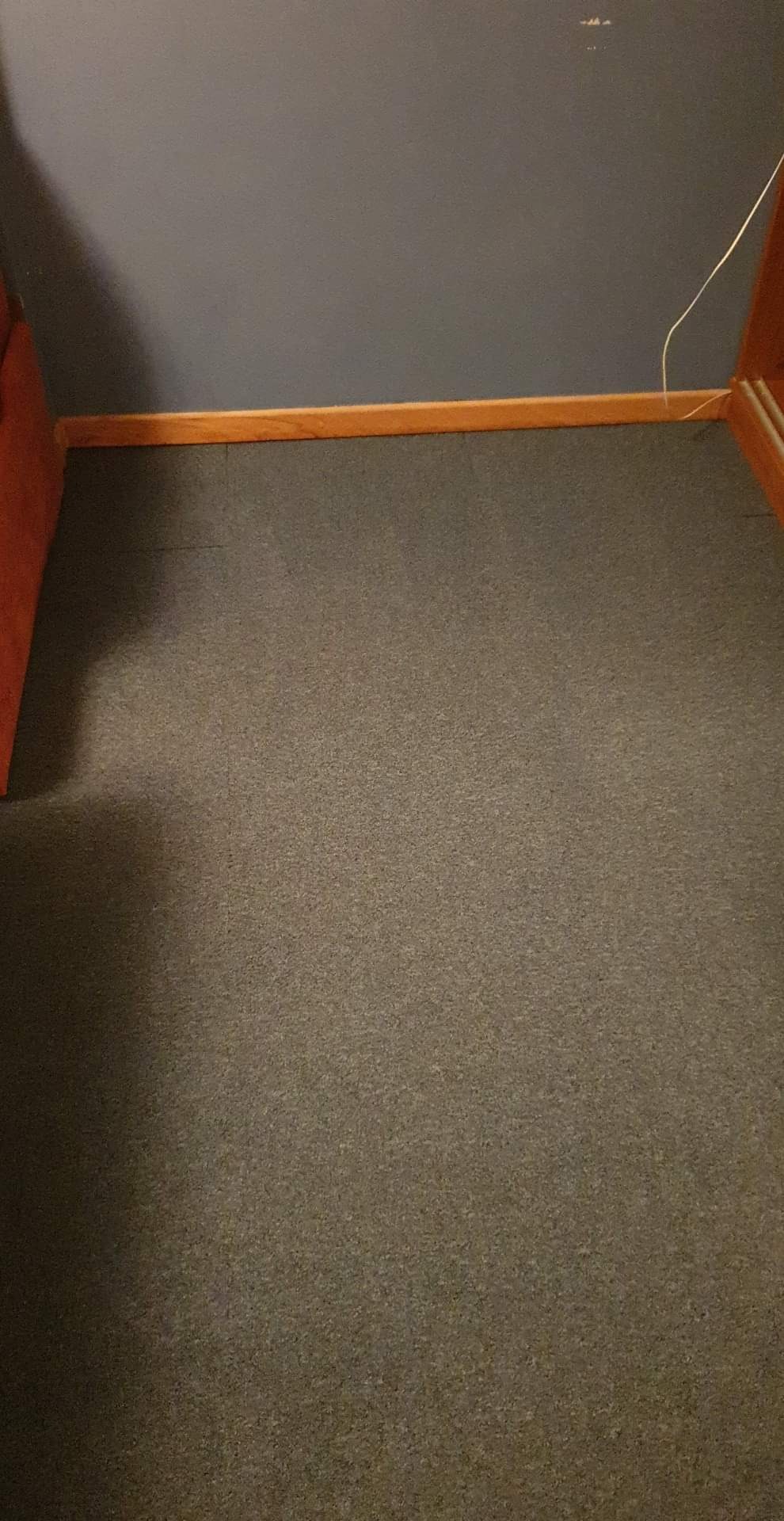 Szary dywan w rogu pokoju z ciemnoszarą ścianą i brązową listwą przypodłogową, widoczny fragment ramy mebla po lewej stronie i kabel elektryczny przy ścianie po prawej stronie.