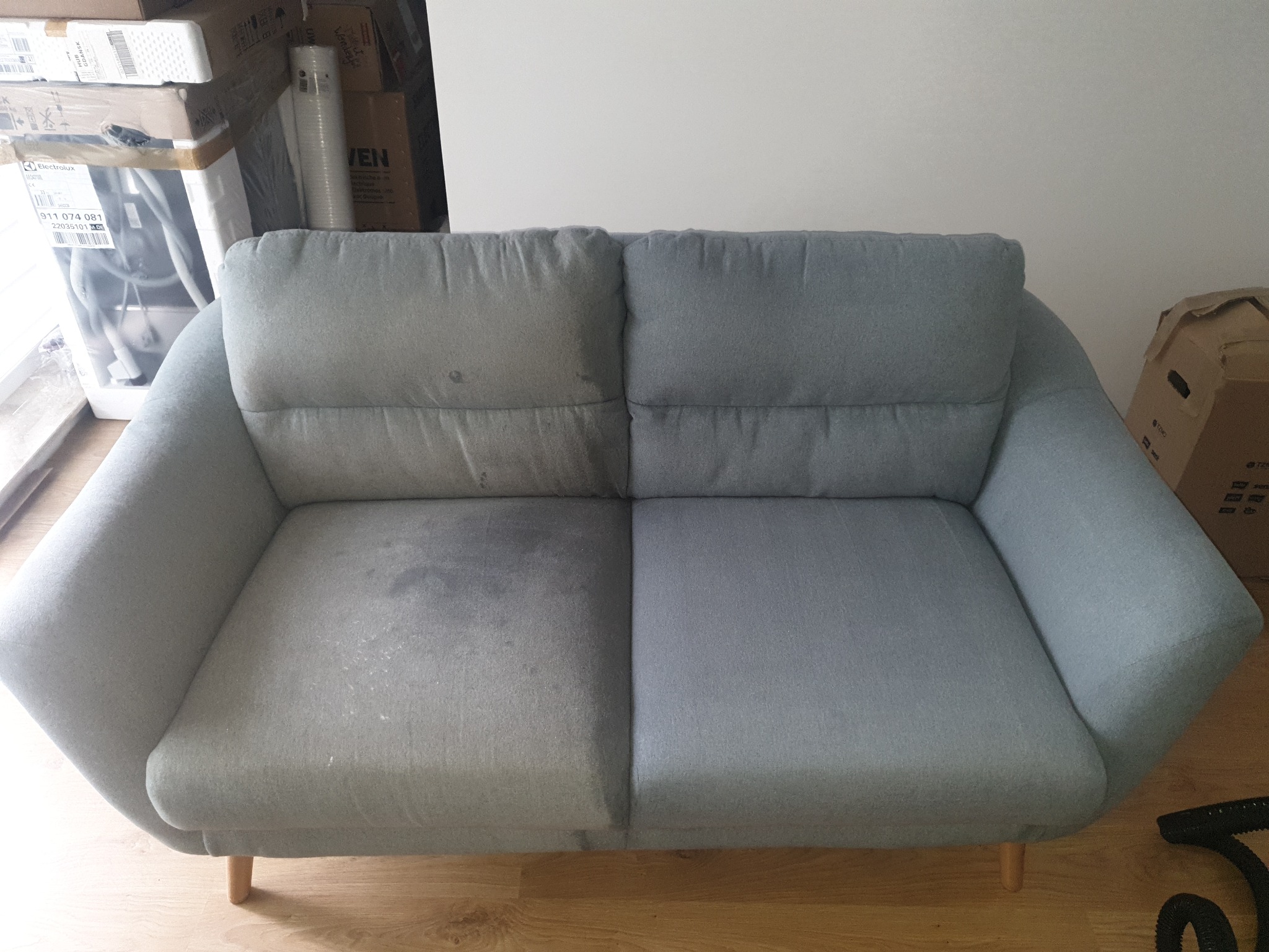 Dwuosobowa kanapa w odcieniach szarości, pokazująca różnicę w wyglądzie przed i po czyszczeniu, z wyraźnie widoczną plamą na lewej połowie i czystą prawą połową. Widoczne pudełka w tle i fragment...