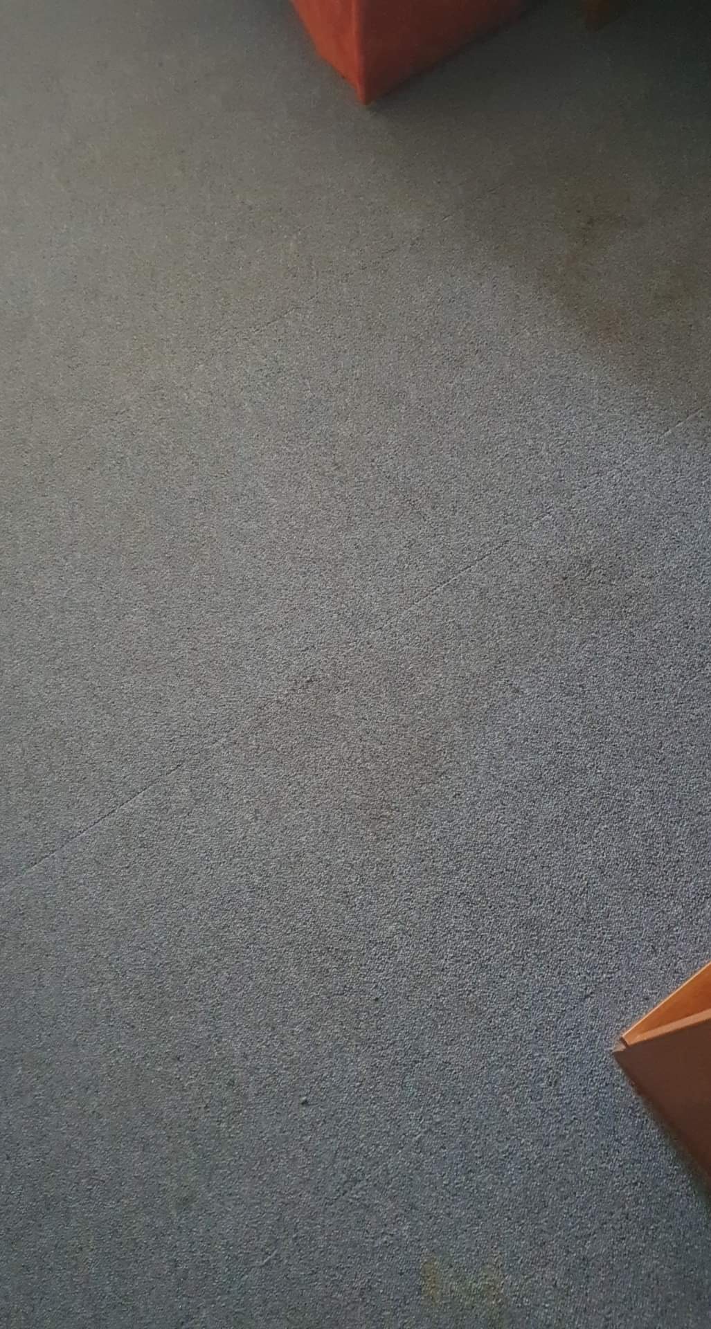 Szary dywan z widocznymi plamami i fragmentem pomarańczowego mebla w rogu.