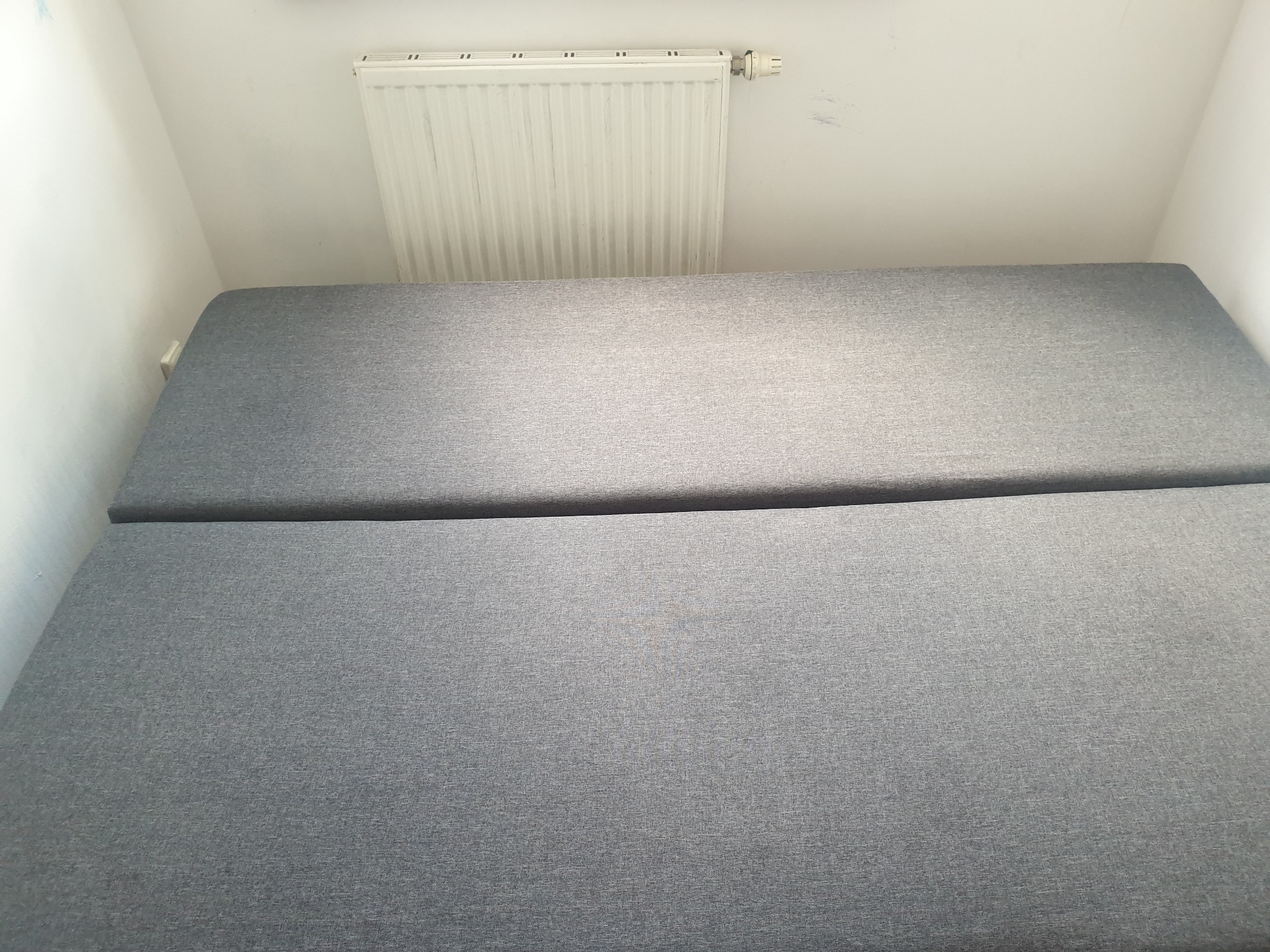 Szary, rozłożony narożnik tapicerowany w rogu pokoju, widoczny grzejnik na ścianie.