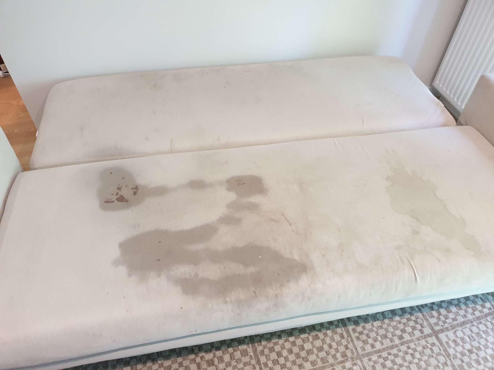 Jasna kanapa z widocznymi, mokrymi plamami po zalaniu lub czyszczeniu, leżąca na wzorzystym dywanie.