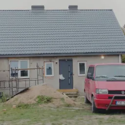 Szary dach z blachy trapezowej na parterowym domu z dwoma kominami, widoczny czerwony samochód dostawczy VW i sterta piasku przed wejściem.