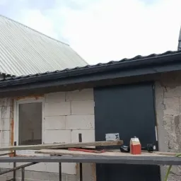 Nowa, czarna rynna zamontowana na budynku z białego bloczka, widoczne narzędzia i materiały budowlane, w tle budynek z dachem z blachy trapezowej.