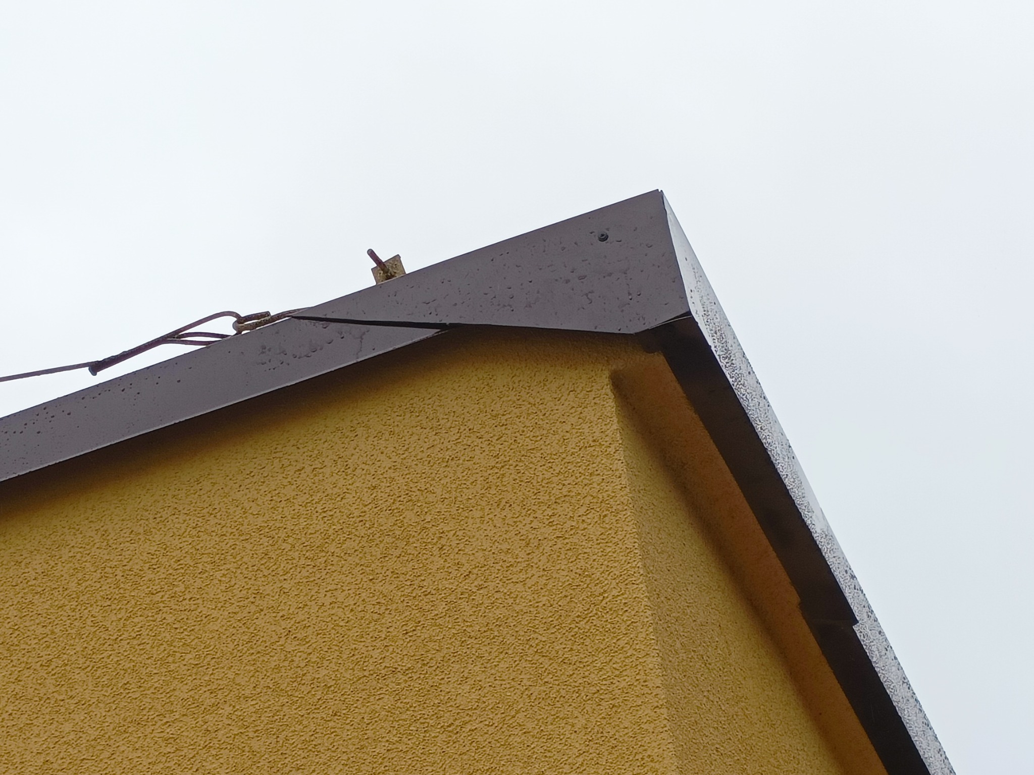 Szczegół nowego, ciemnobrązowego orynnowania na krawędzi dachu budynku z żółtą elewacją o strukturze baranka, widoczny fragment instalacji odgromowej.