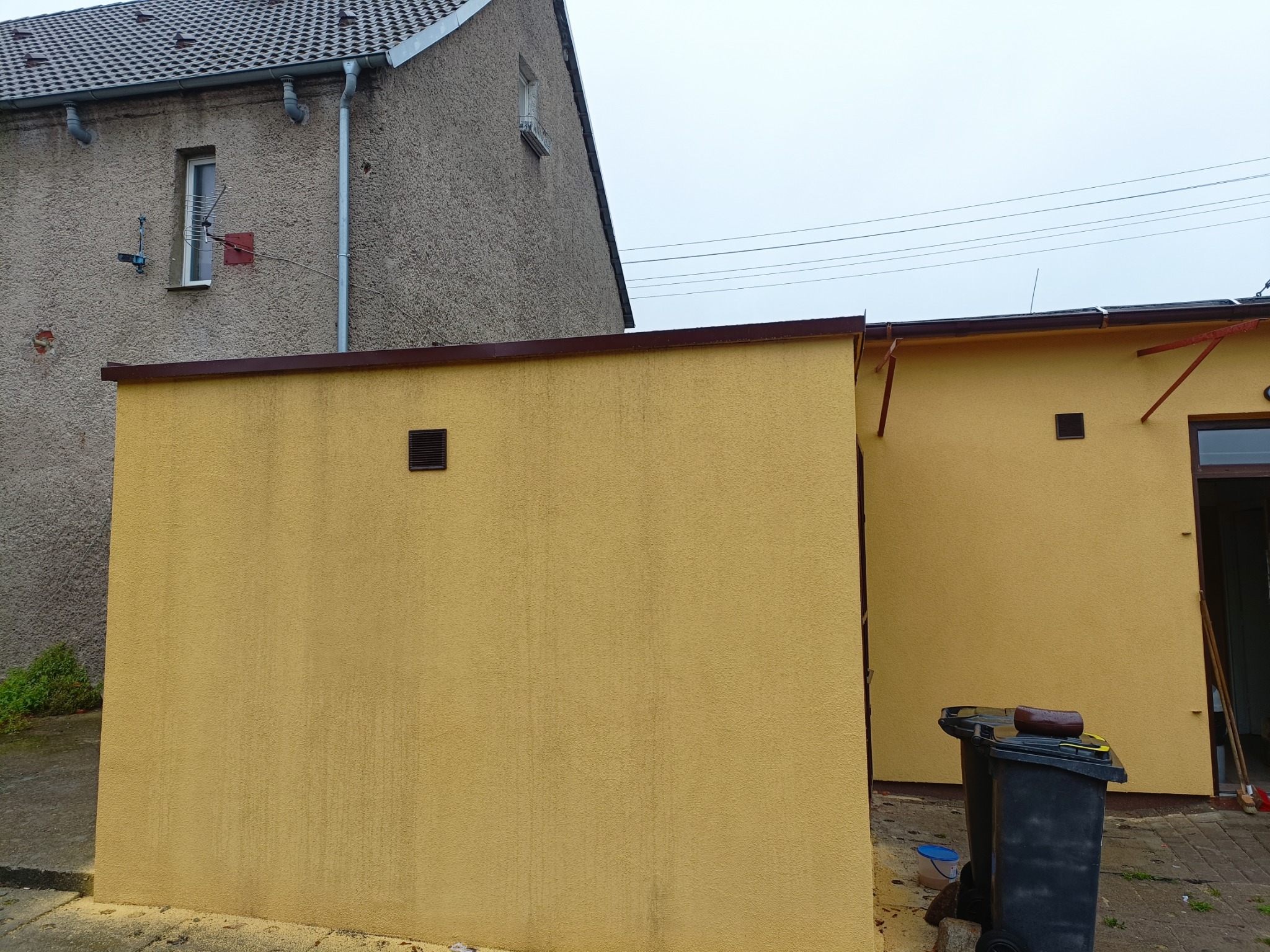Żółta ściana garażu z brązowym wykończeniem, widoczny fragment elewacji domu z anteną i rynnami, dwa czarne pojemniki na śmieci, pochmurne niebo.