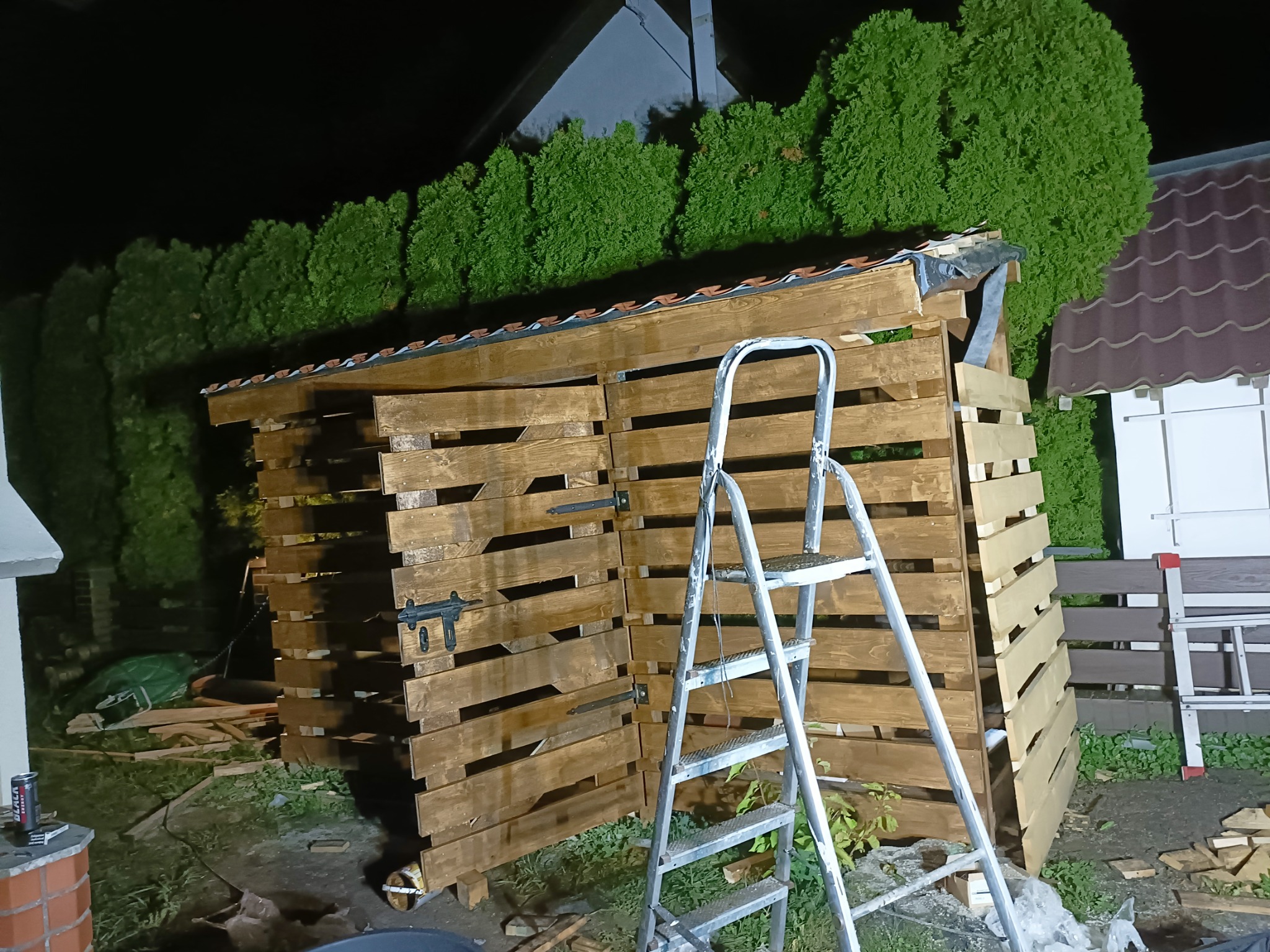 Drewniana wiata na drewno z zadaszeniem z dachówki, częściowo otwarta, z drabiną przystawioną z przodu na zewnątrz w nocy.