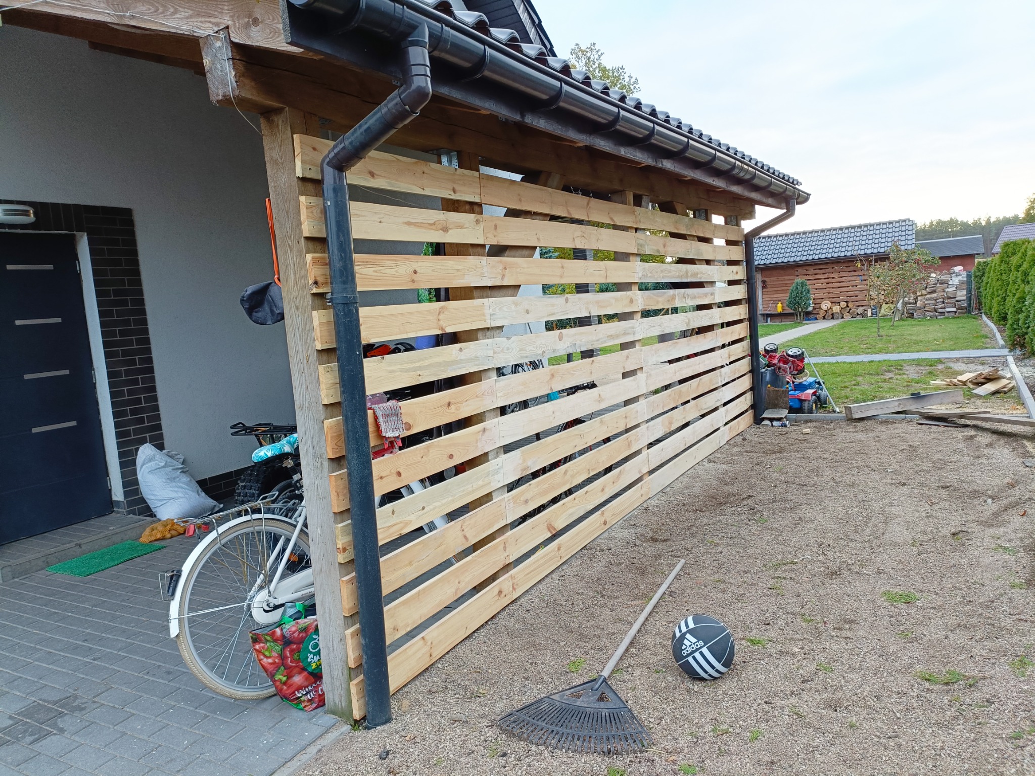 Zadaszona wiata z drewnianą ażurową ścianą z desek, widoczne rynny, rower oparty o ścianę, grabie i piłka na żwirowym podłożu.