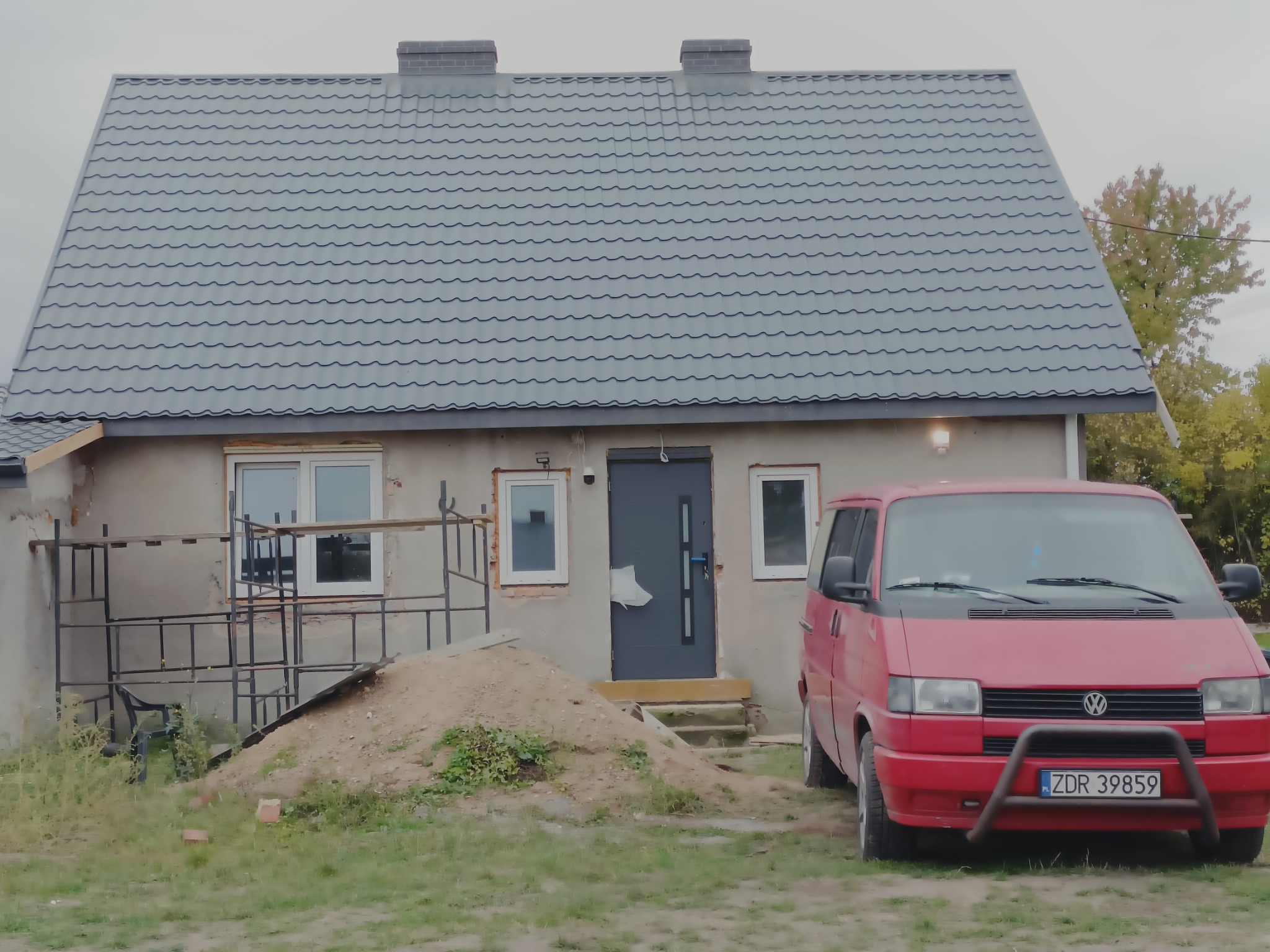 Szary dach z blachy trapezowej na parterowym domu z dwoma kominami, widoczny czerwony samochód dostawczy VW i sterta piasku przed wejściem.