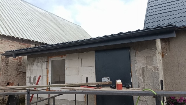 Nowa, czarna rynna zamontowana na budynku z białego bloczka, widoczne narzędzia i materiały budowlane, w tle budynek z dachem z blachy trapezowej.