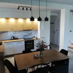 Nowoczesna kuchnia z białymi i drewnianymi frontami szafek, blatem imitującym marmur, drewnianym stołem z czarnymi krzesłami i schodami z podświetleniem w tle.