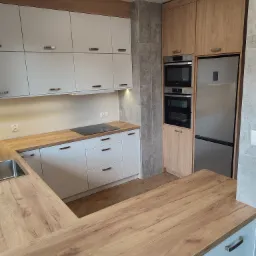 Nowoczesna kuchnia: białe szafki z drewnianymi blatami i akcentami, zabudowany sprzęt AGD, szara ściana, widok na wyspę kuchenną.