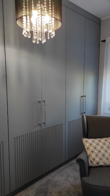 Szara szafa wnękowa z frezowanymi pionowymi zdobieniami na dolnych frontach, długie kryształowe uchwyty, kryształowy żyrandol w tle, fotel z poduszką w geometryczny wzór.