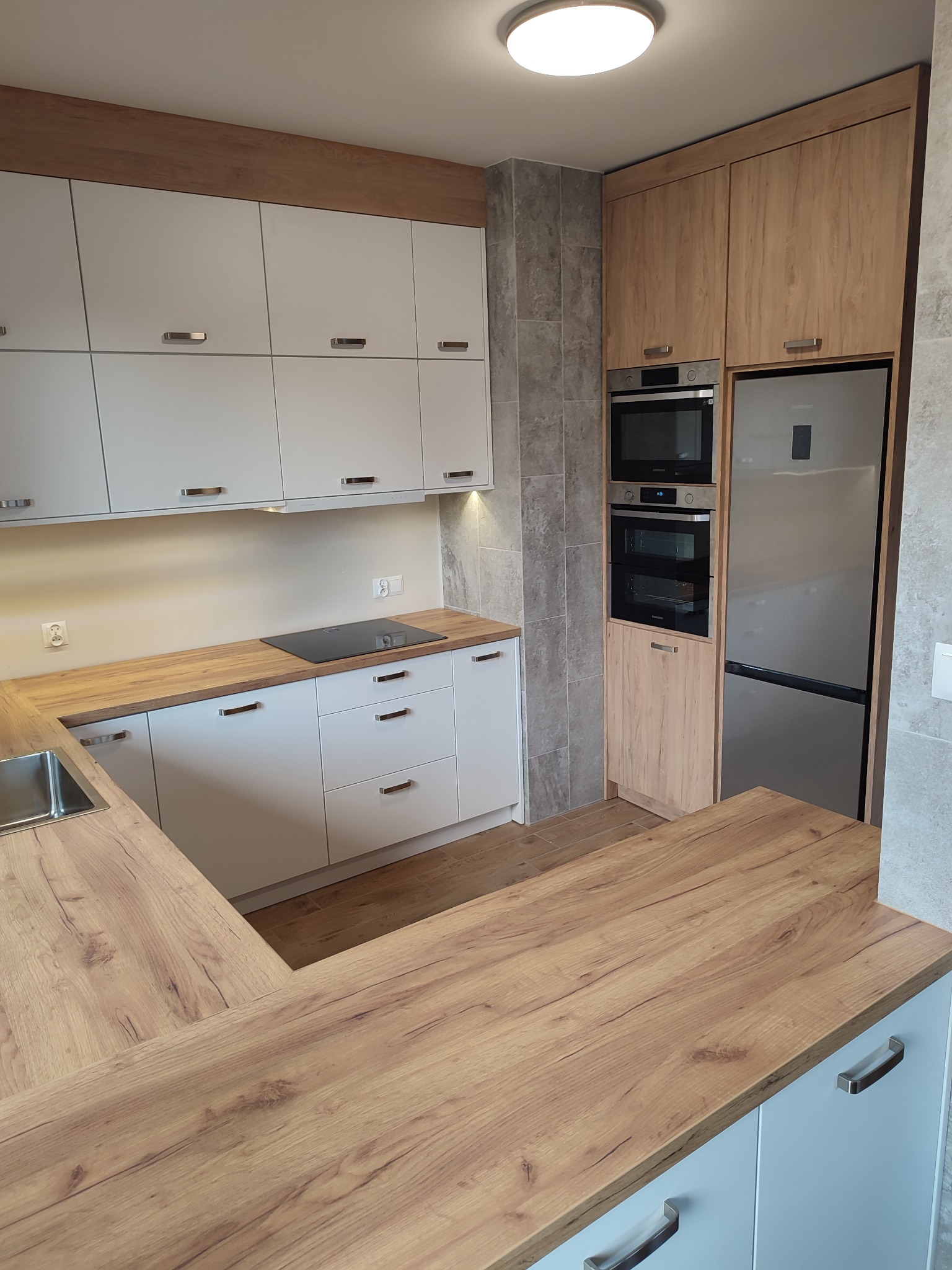 Nowoczesna kuchnia: białe szafki z drewnianymi blatami i akcentami, zabudowany sprzęt AGD, szara ściana, widok na wyspę kuchenną.