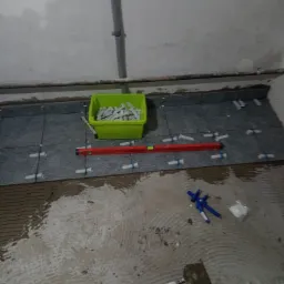 Układanie szarych płytek podłogowych z użyciem krzyżyk&oacute;w dystansowych i poziomicy; widoczny zielony pojemnik z zapasem krzyżyk&oacute;w i ślady zaprawy na podłodze.