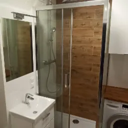 Nowoczesna łazienka z kabiną prysznicową typu walk-in, umywalką z szafką, lustrem, pralką i drewnopodobnymi płytkami na ścianie kabiny oraz podłodze.