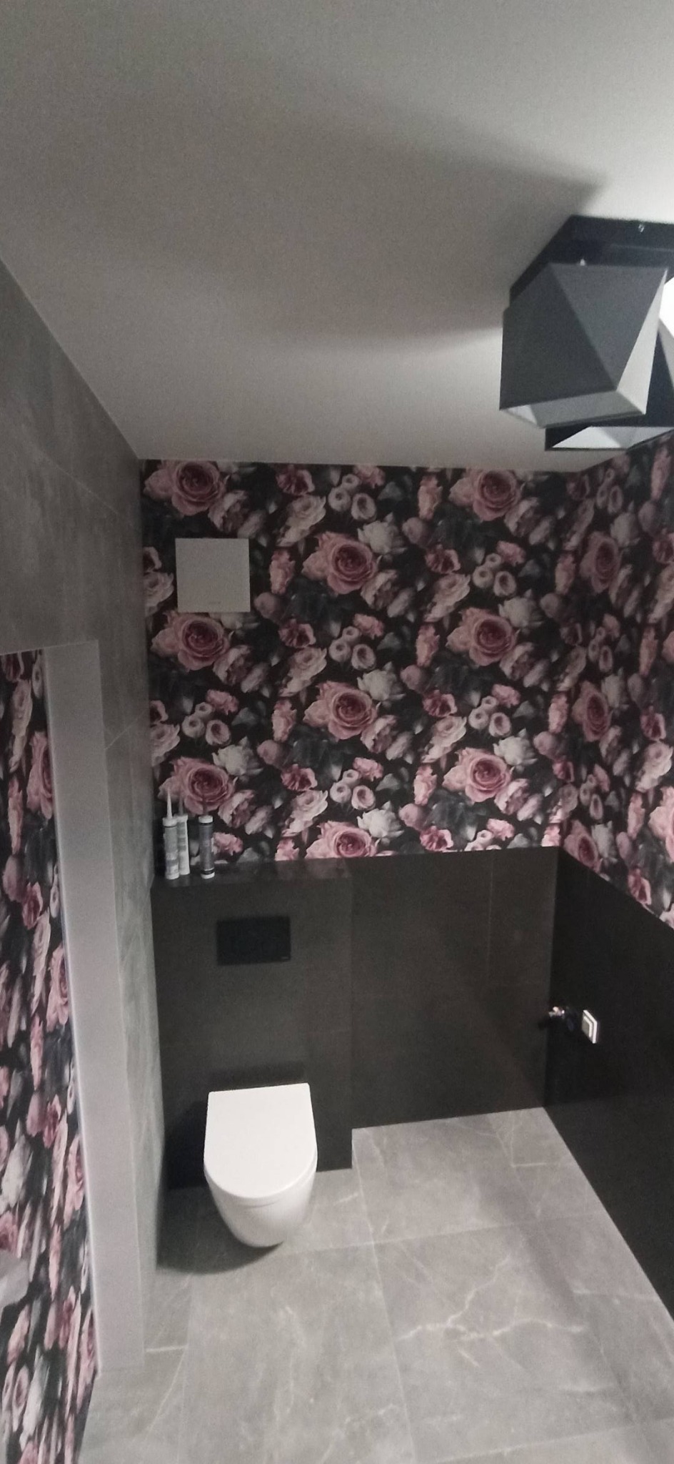 Nowoczesna łazienka z wiszącą toaletą, szarą armaturą i tapetą w kwiaty, oświetlona geometryczną lampą sufitową.