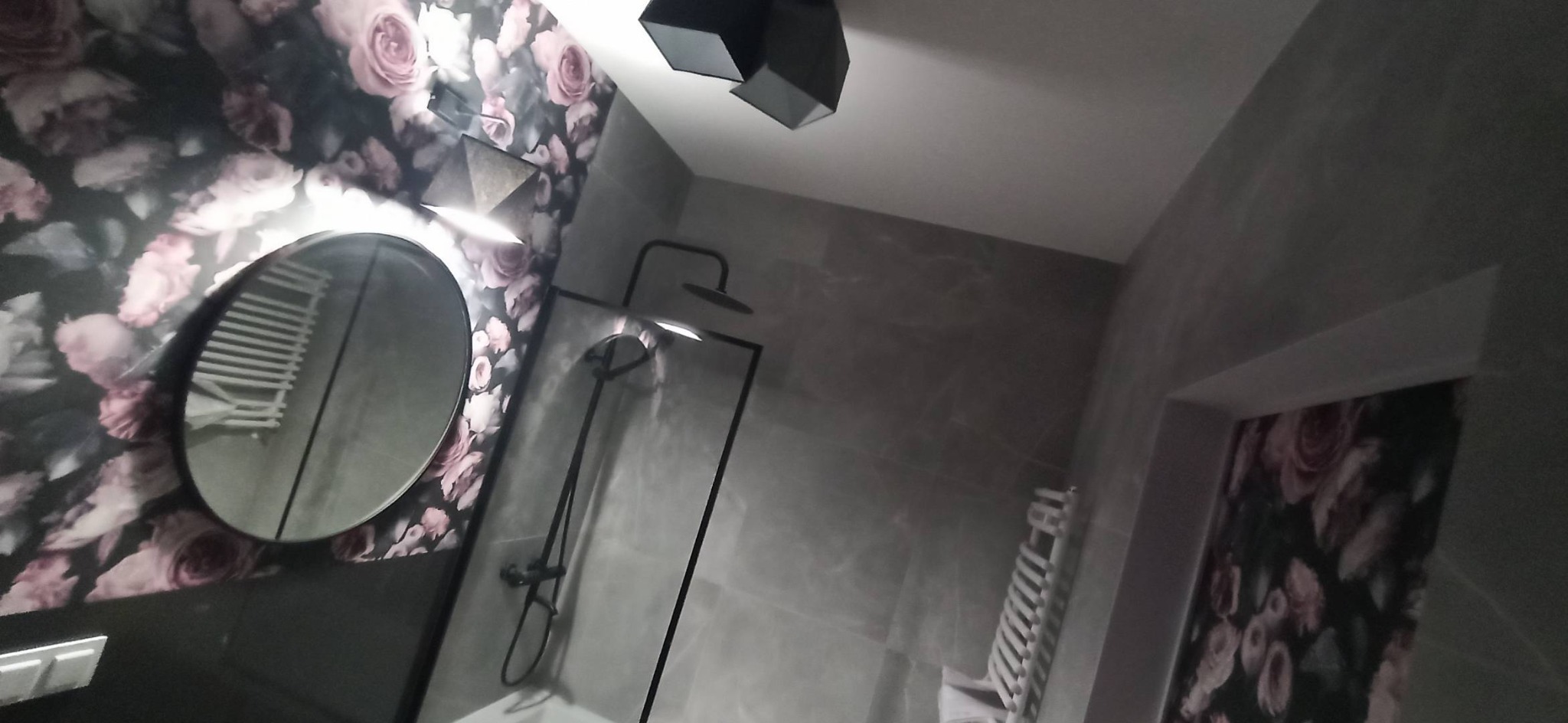 Nowoczesna łazienka z szarą armaturą, kabiną prysznicową typu walk-in, okrągłym lustrem i tapetą w kwiaty.