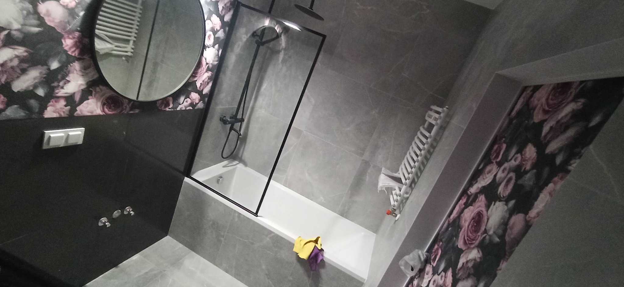 Wykończona łazienka w Łodzi z wanną za czarną szybą, szare płytki imitujące marmur, ściana z tapetą w kwiaty i okrągłe lustro.
