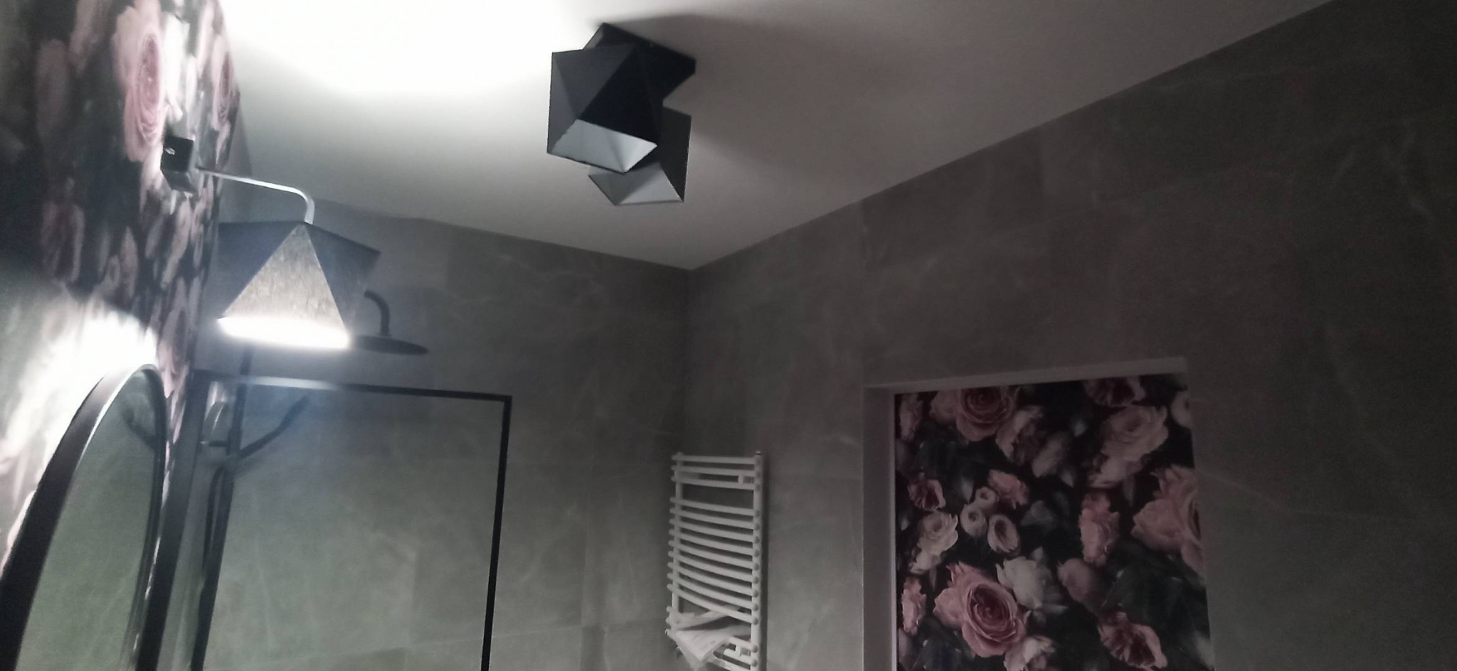 Nowoczesna łazienka z szarą, marmurkową ścianą, geometryczną lampą sufitową, grzejnikiem drabinkowym i fragmentem lustra z oświetleniem punktowym.