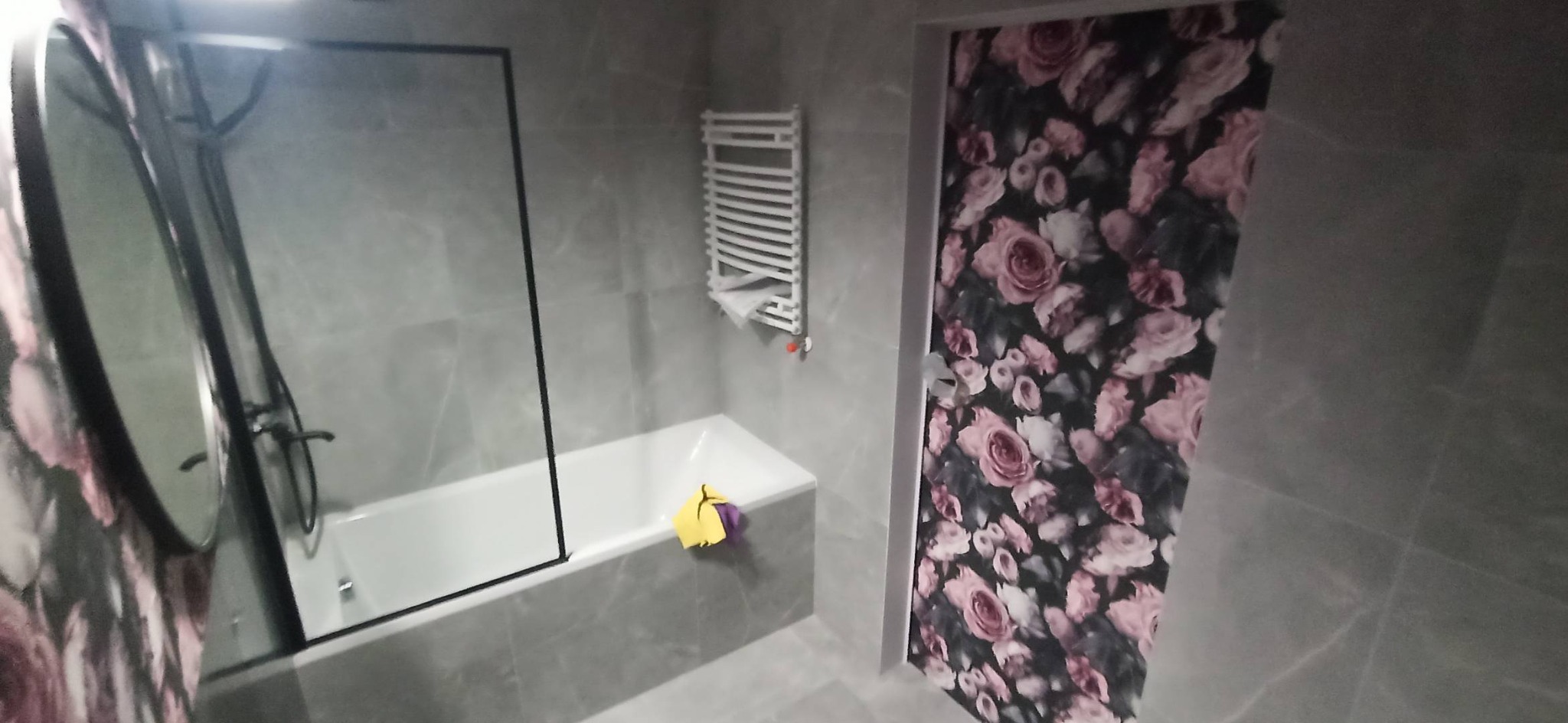 Wykończona łazienka z szarymi płytkami imitującymi marmur, białą wanną z czarną ramą kabiny prysznicowej, grzejnikiem drabinkowym i drzwiami z motywem różanych kwiatów.