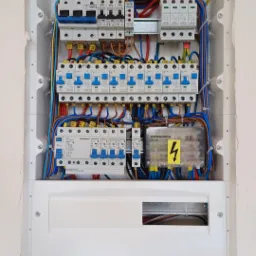 Rozdzielnica elektryczna w białej obudowie, widoczne bezpieczniki, przewody w różnych kolorach oraz listwa zaciskowa, zamontowana w ścianie.