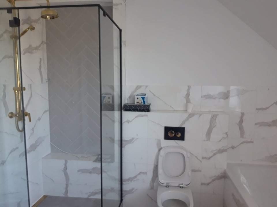 Nowoczesna łazienka na poddaszu z kabiną prysznicową typu walk-in z czarną ramą, złotą armaturą i płytkami imitującymi marmur ułożonymi w jodełkę wewnątrz kabiny.