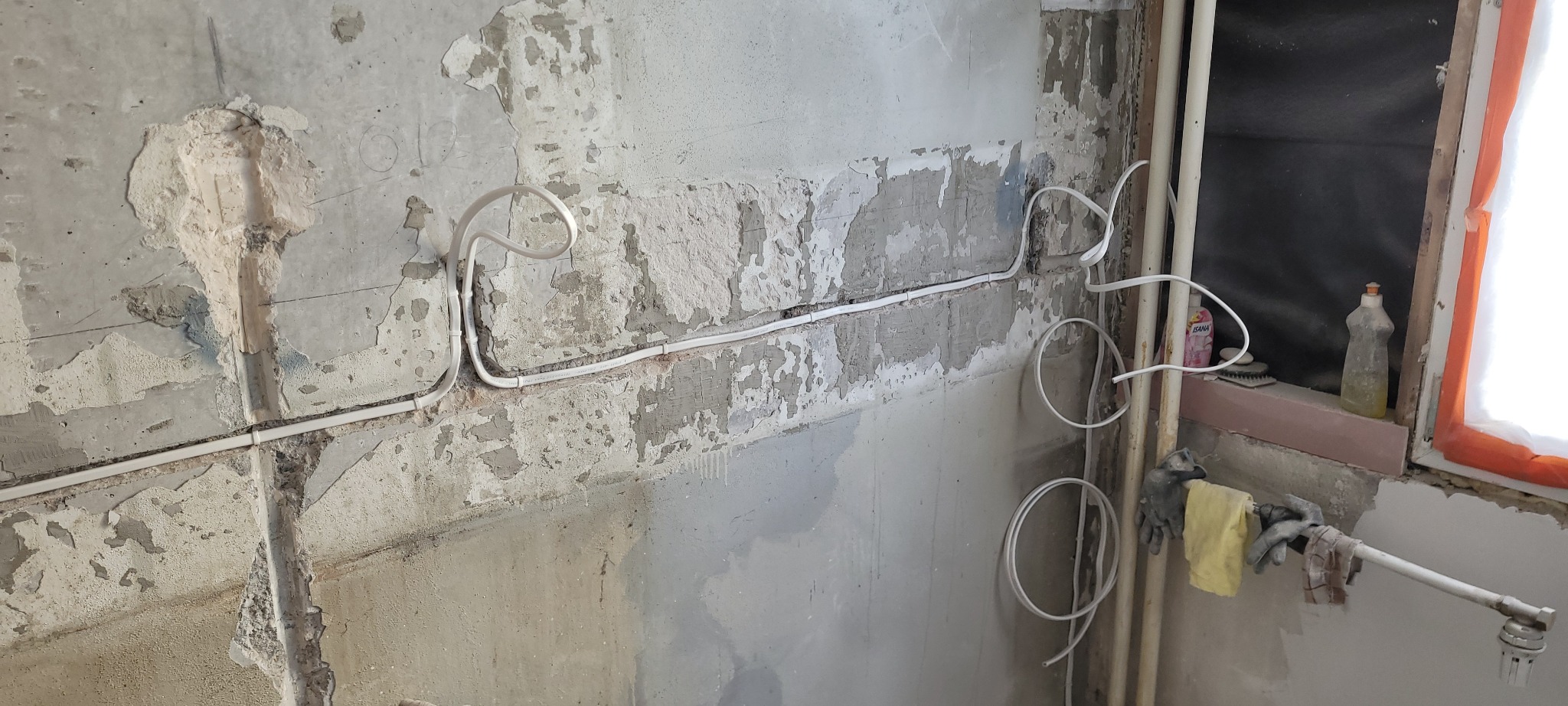 Ściana w trakcie remontu z widocznymi przewodami elektrycznymi, fragmentami starego tynku i instalacją hydrauliczną, przygotowana do dalszych prac wykończeniowych.