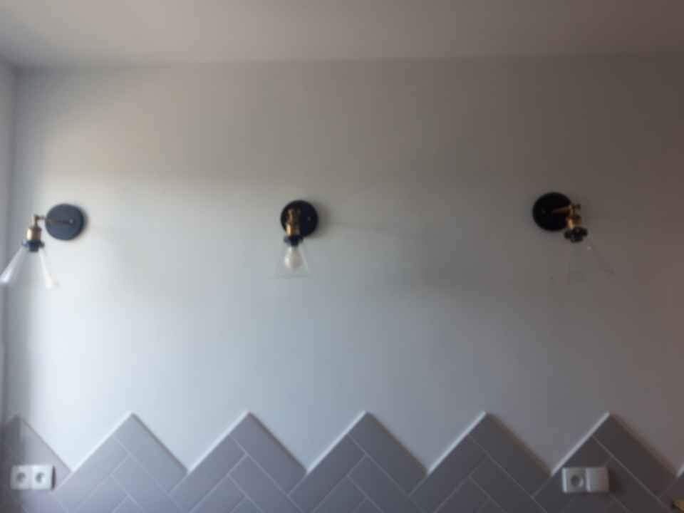 Ściana z szarymi płytkami ułożonymi w jodełkę i trzema lampami ściennymi w stylu industrialnym z żarówkami w kształcie kropli.