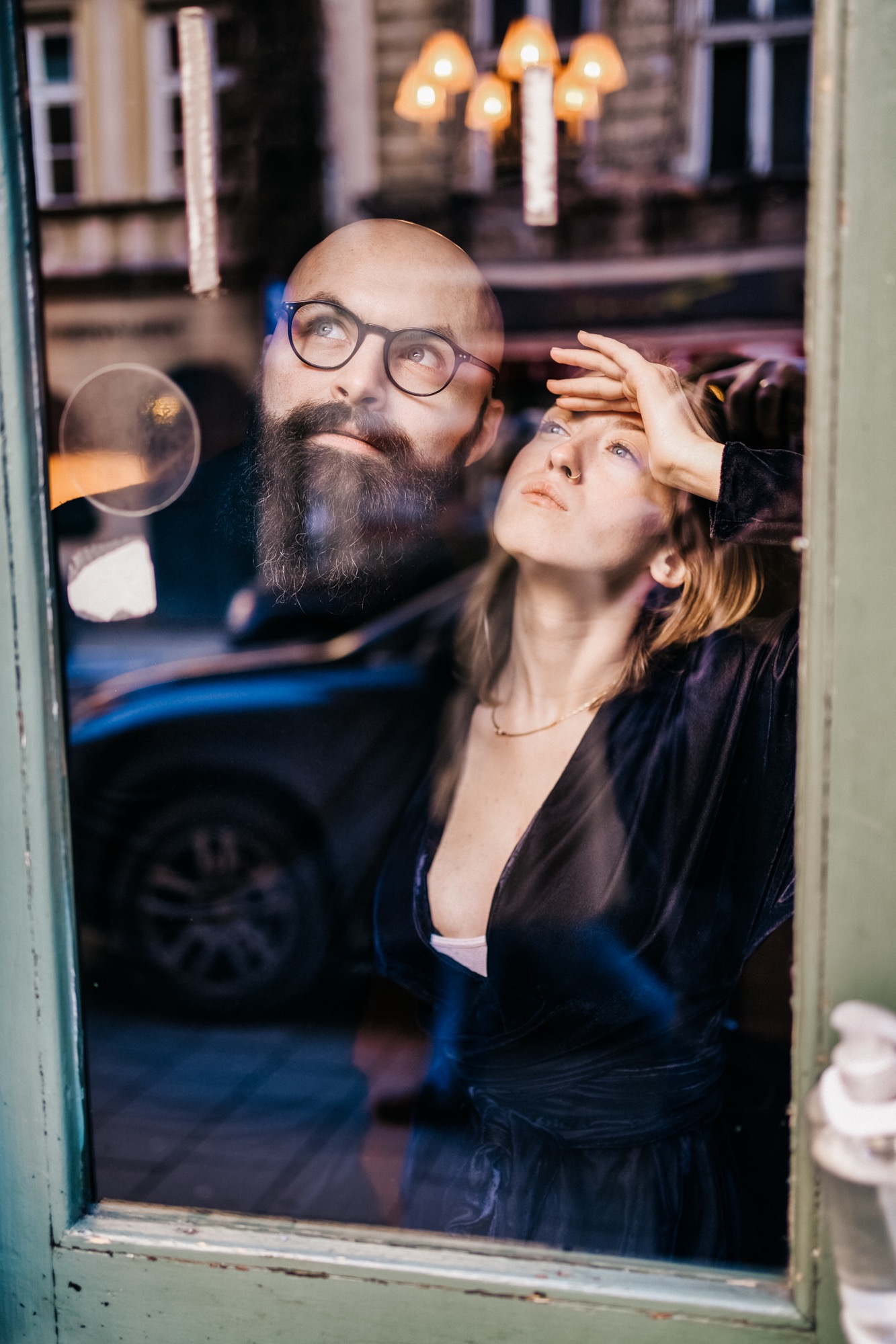 Portret pary przez szybę: mężczyzna z brodą i okularami patrzy w górę, kobieta w welurowej sukience opiera dłoń na czole, w tle odbicie ulicy i lamp