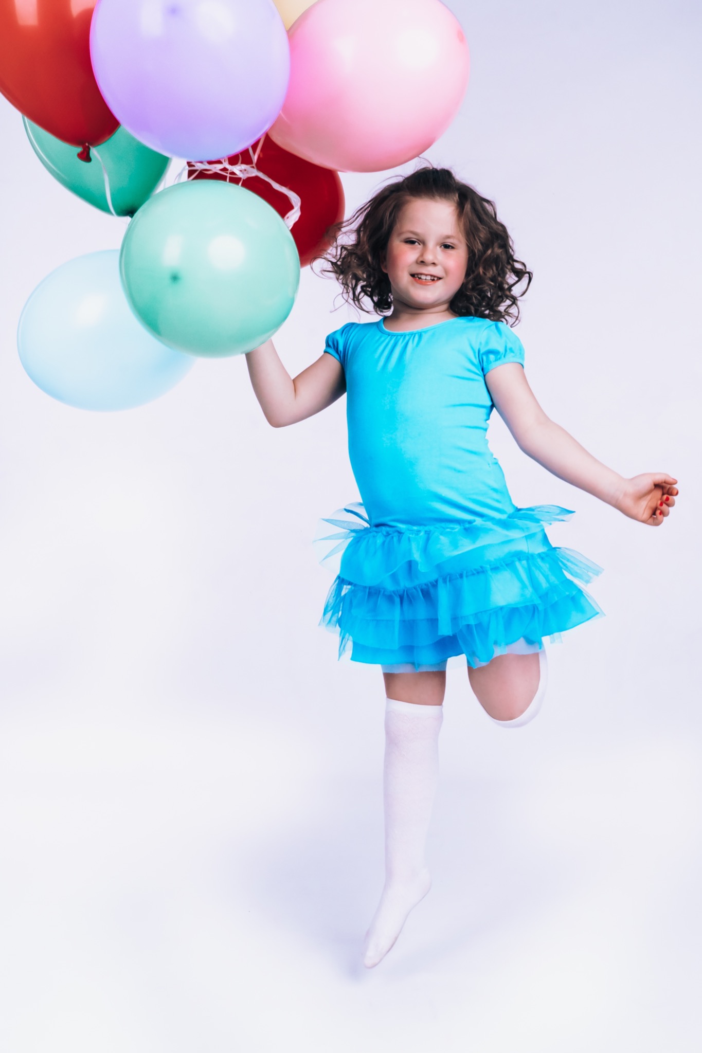 Uśmiechnięta dziewczynka w błękitnej sukience baletnicy skacze trzymając pęk kolorowych balonów na białym tle.