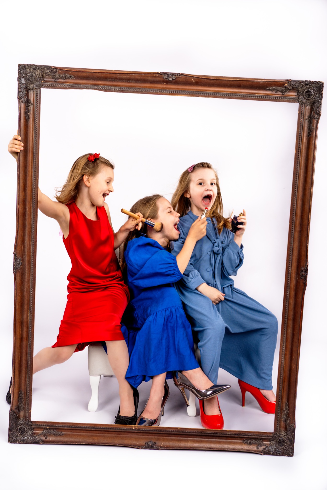 Trzy dziewczynki w eleganckich sukienkach i butach na obcasie bawią się makijażem, pozując wewnątrz dużej, ozdobnej ramy na białym tle.