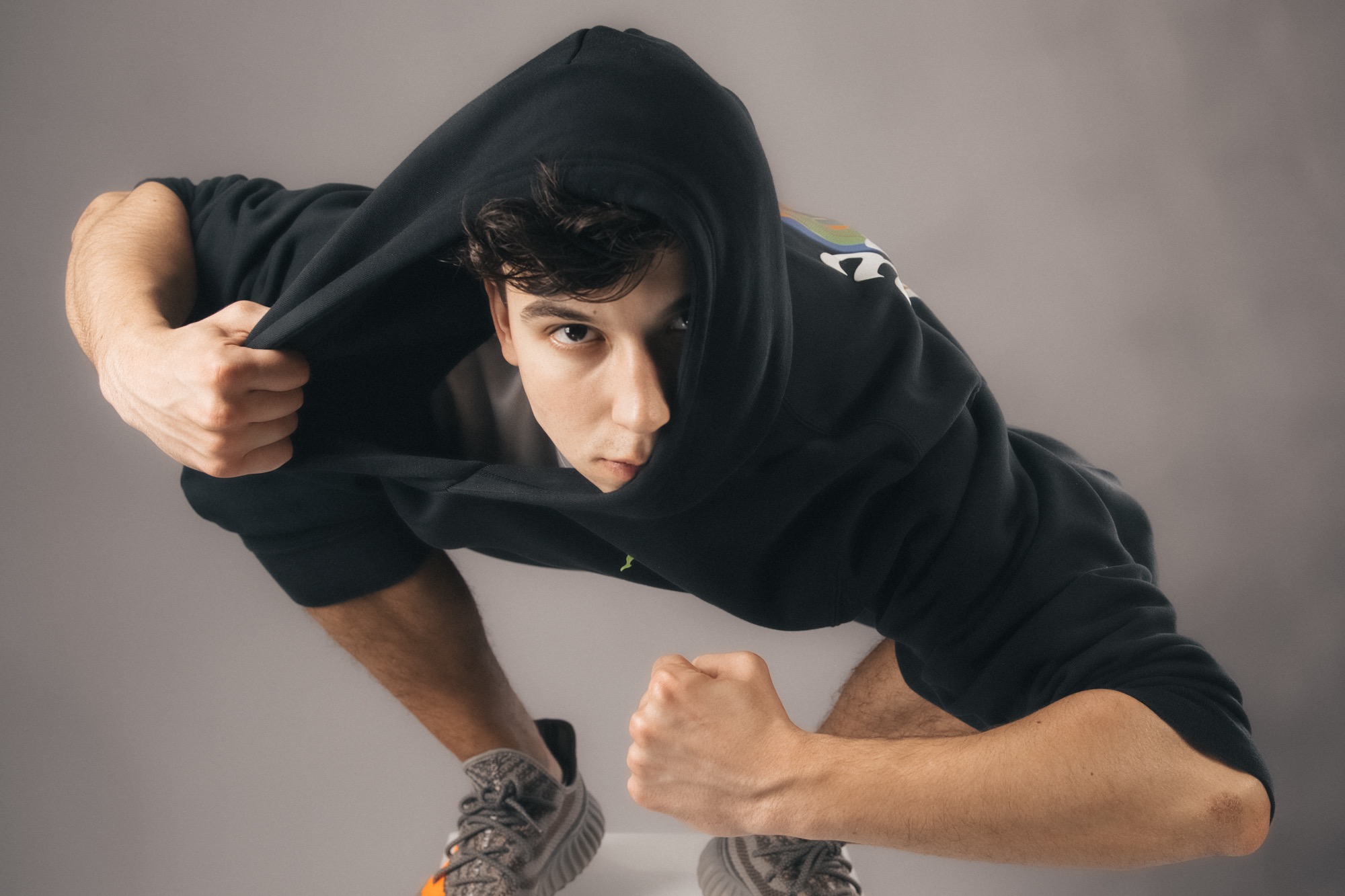 Młody mężczyzna w czarnej bluzie z kapturem, częściowo zakrywającym twarz, patrzy w dół, dłonie zaciśnięte w pięści, widoczne szare buty sportowe z pomarańczowymi akcentami na tle szarego tła.