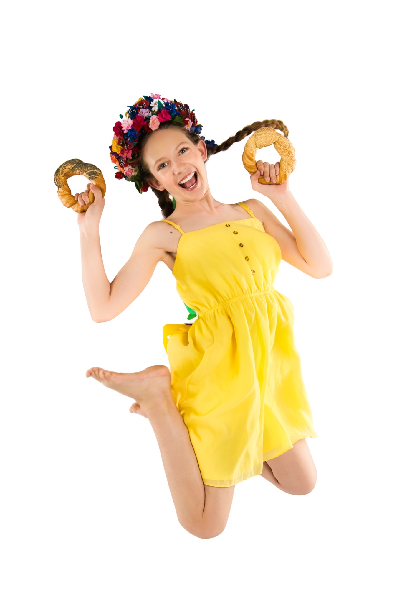 Uśmiechnięta dziewczyna w żółtej sukience i wianku na głowie, skacze z radości trzymając w dłoniach dwa precle z różnymi posypkami na białym tle.