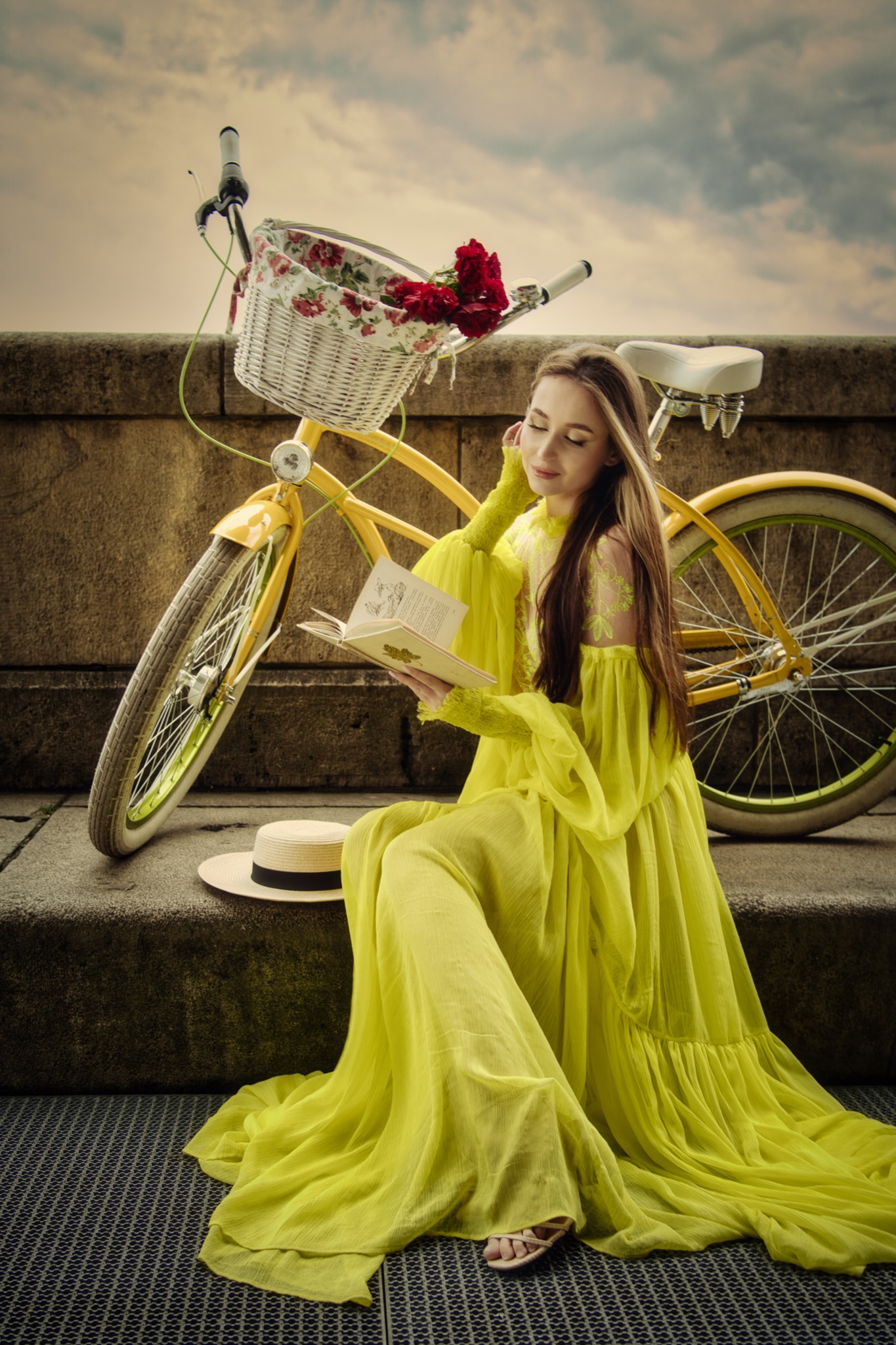 Kobieta w długiej, zwiewnej, cytrynowej sukni siedzi na schodach z książką, obok żółty rower z kwiatami w koszyku i kapelusz.