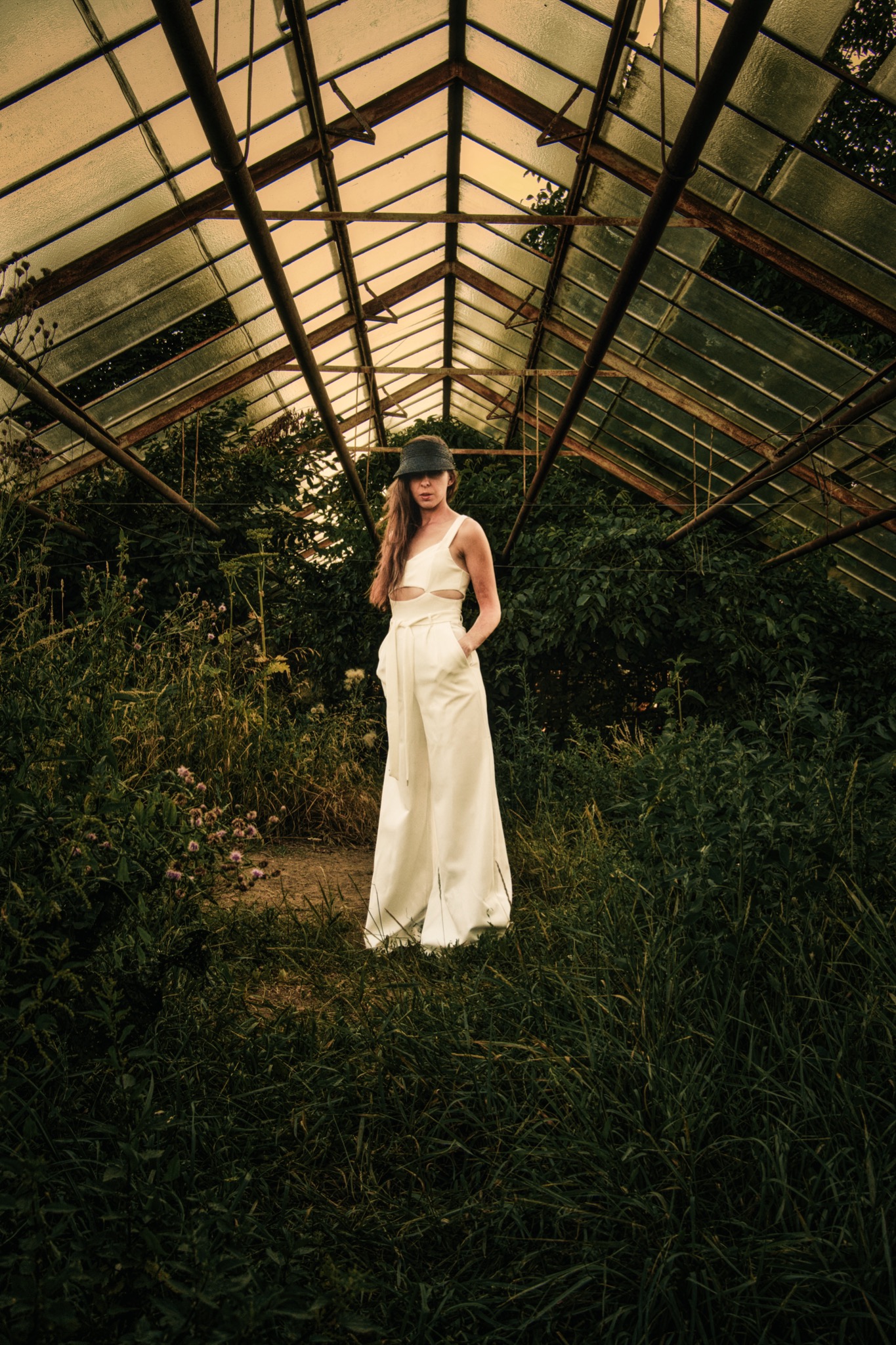 Kobieta w białym kombinezonie i kapeluszu stoi w opuszczonej szklarni, otoczona bujną zielenią i zardzewiałą konstrukcją dachu.