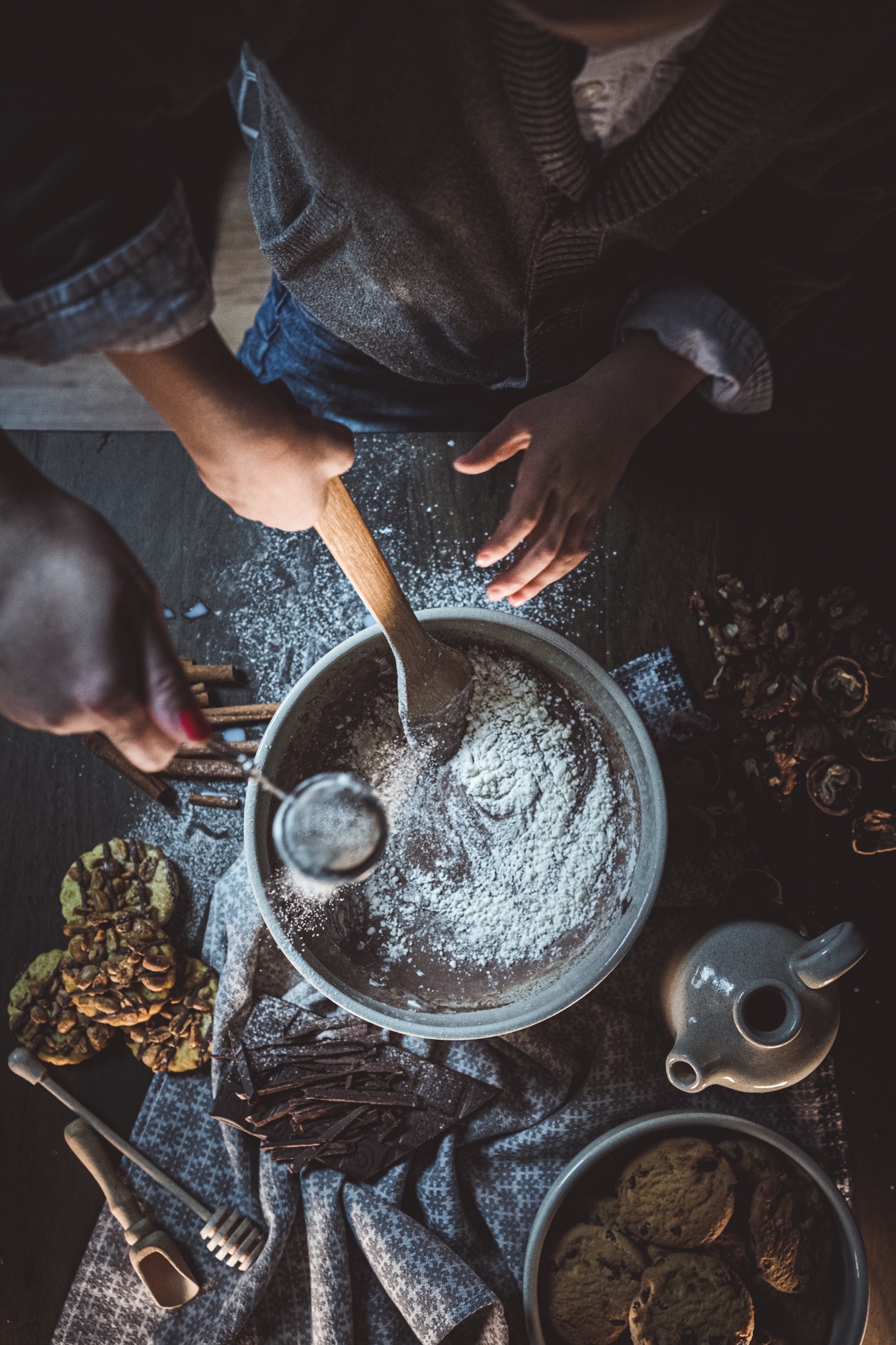 Przygotowywanie ciasta czekoladowego: przesiewanie mąki przez sitko do miski z masą, obok kawałki czekolady, ciasteczka, orzechy włoskie i drewniane przybory kuchenne, widok z góry w ciemnym...