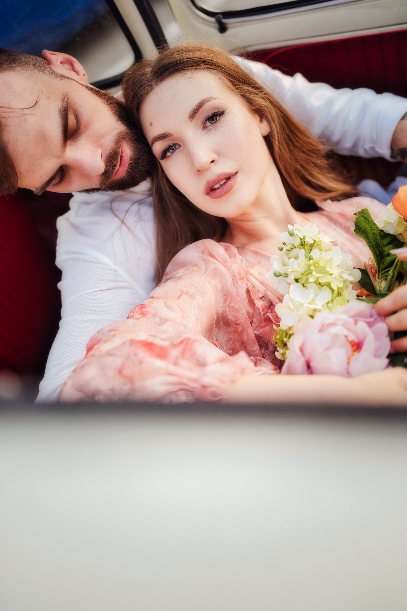 Kobieta w zwiewnej sukience trzyma bukiet kwiatów, opierając się na ramieniu mężczyzny w białej koszuli, wnętrze samochodu z czerwoną tapicerką, delikatne oświetlenie.