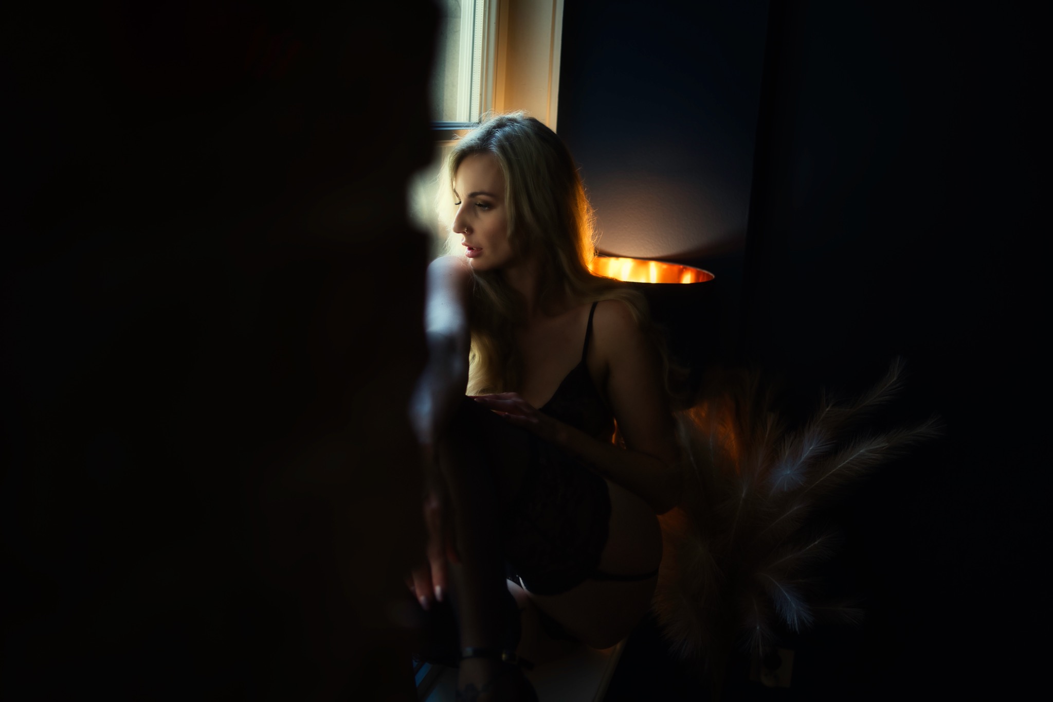 Kobieta w bieliźnie i pończochach siedzi przy oknie, oświetlona ciepłym światłem, z piórami dekoracyjnymi obok.
