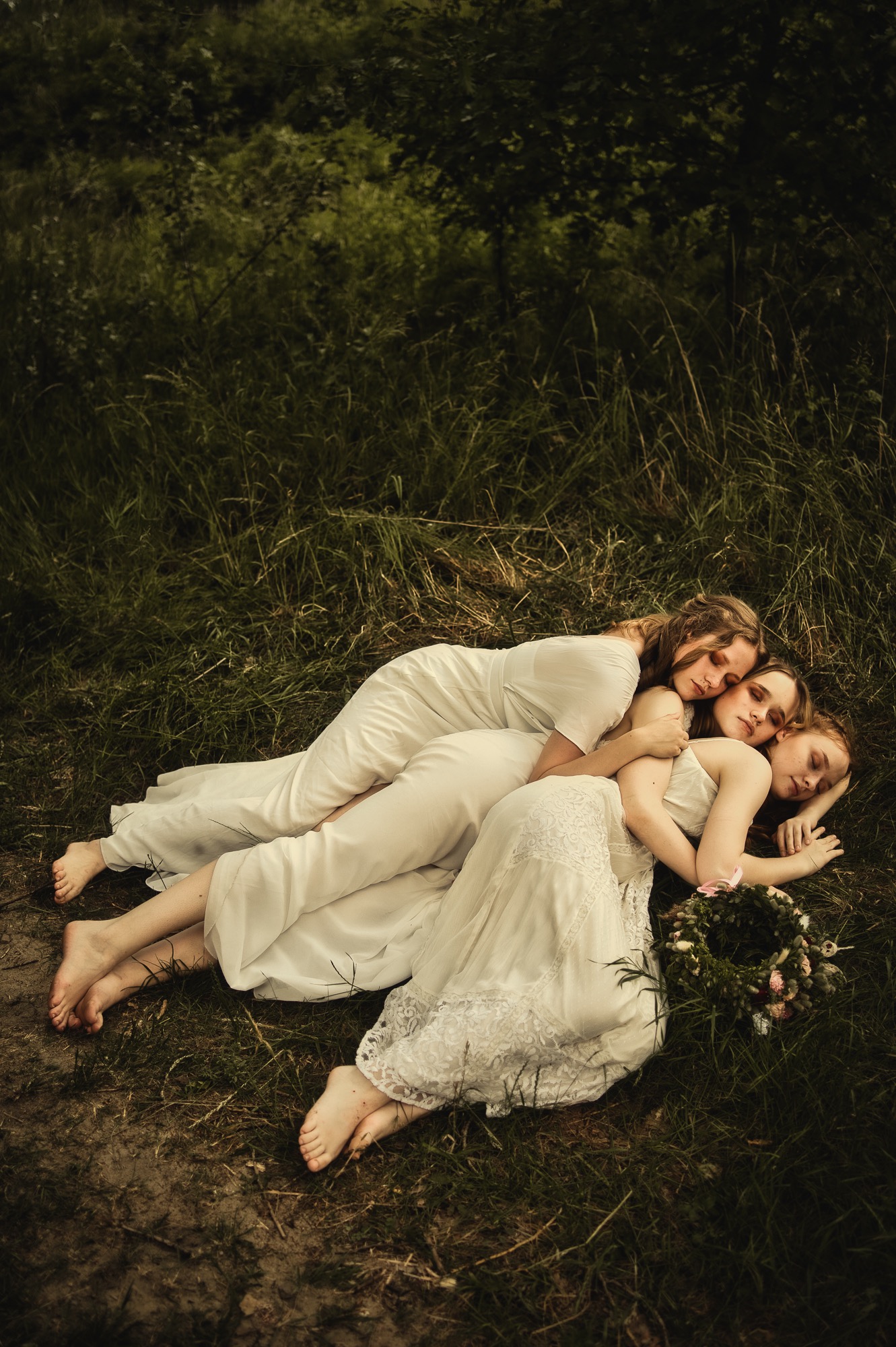 Trzy kobiety w zwiewnych białych sukienkach leżące na trawie z wiankiem z kwiatów, w naturalnym oświetleniu, tworząc intymną i artystyczną kompozycję.