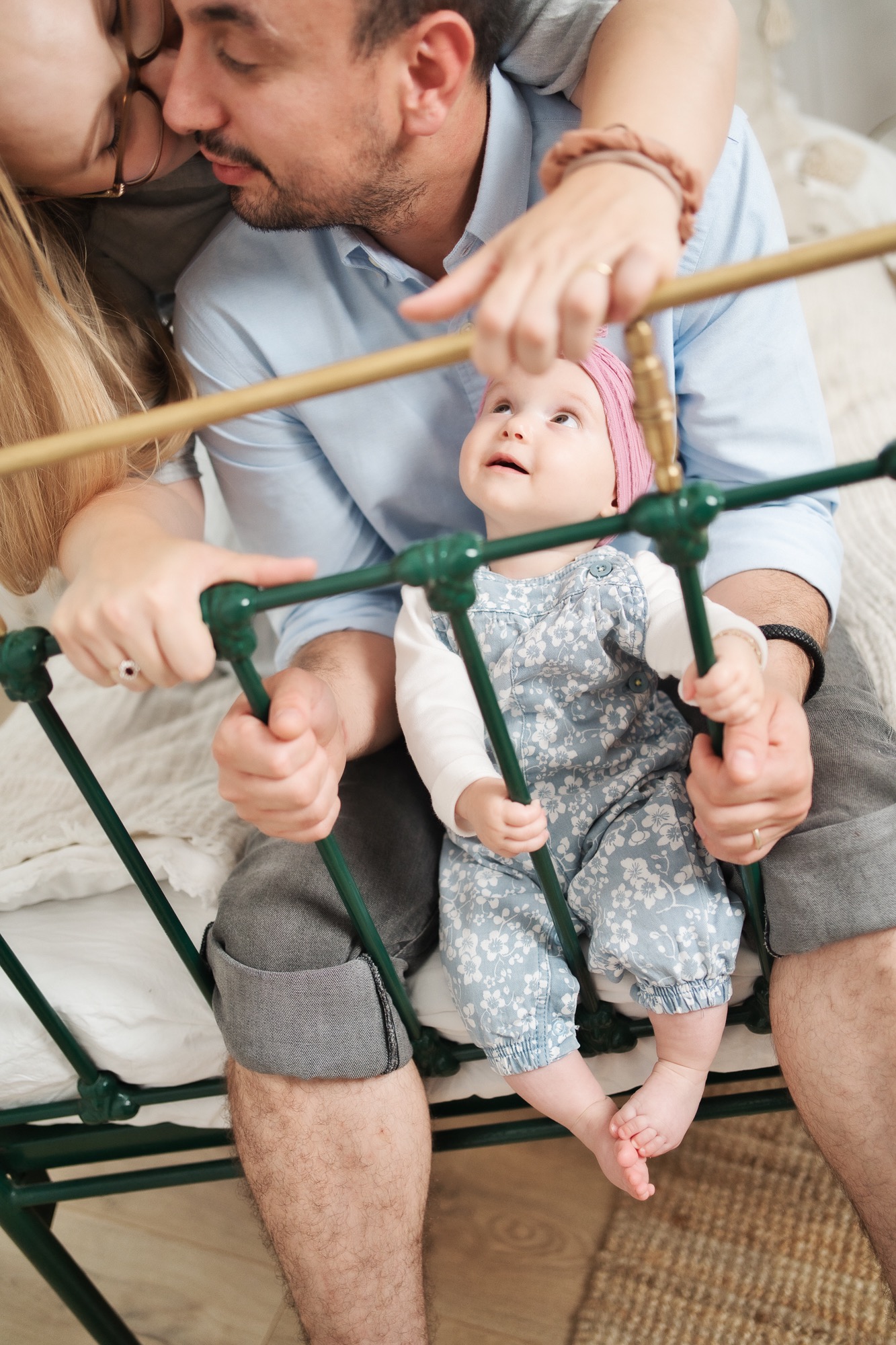 Rodzice trzymają małą dziewczynkę w kwiecistym kombinezonie i różowej czapeczce, stojącą na zielonym łóżku z metalową ramą, z uśmiechem spoglądającą w górę.