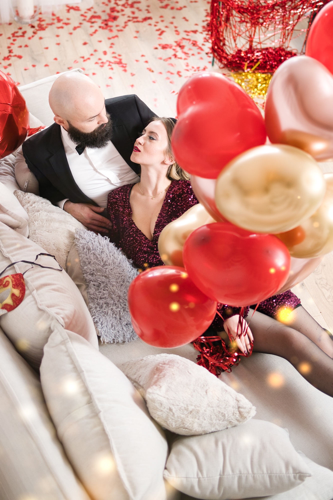 Elegancka para na kanapie, mężczyzna w smokingu, kobieta w cekinowej sukience, otoczeni czerwonymi i złotymi balonami w kształcie serc, konfetti na podłodze.