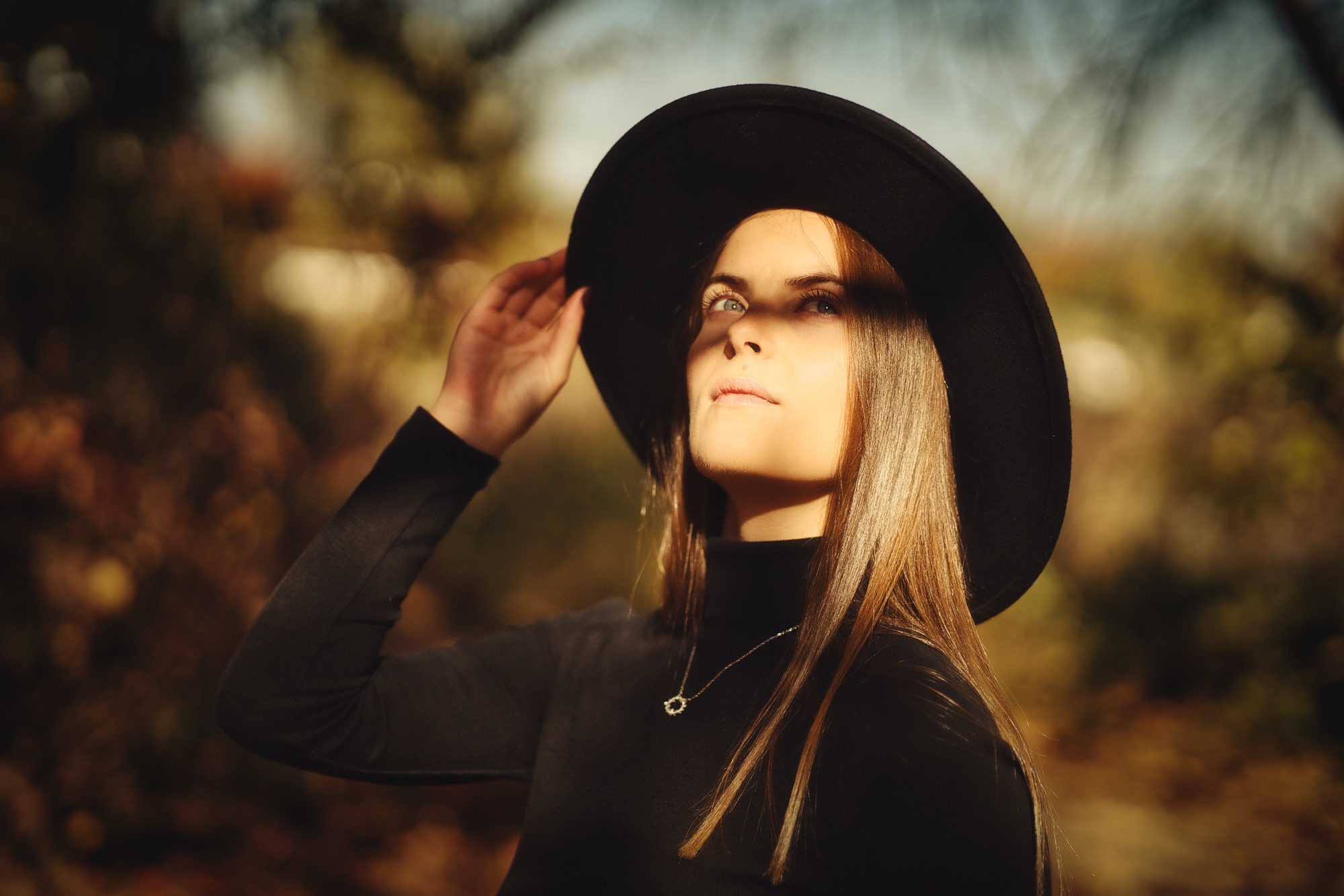 Portret kobiety w czarnym kapeluszu z szerokim rondem, spoglądającej w górę w promieniach słońca, z delikatnym naszyjnikiem na szyi.