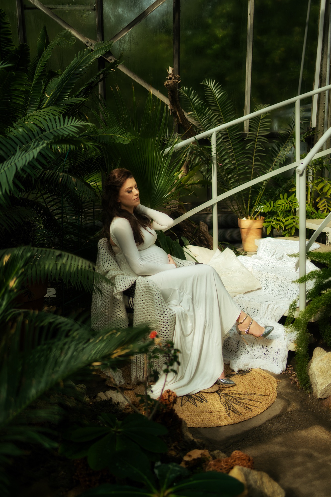 Kobieta w białej sukni siedzi na krześle w otoczeniu tropikalnej roślinności w szklarni, srebrne buty na wysokim obcasie, ręka na brzuchu, zamyślony wyraz twarzy.